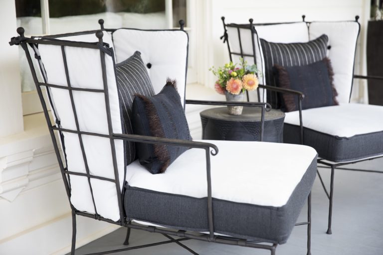 Two wrought iron chairs with white cushions and dark throw pillows are positioned on a patio. A small round table with a vase of fresh flowers sits between them. The setting is outdoors, in front of a window, creating a cozy and inviting atmosphere.